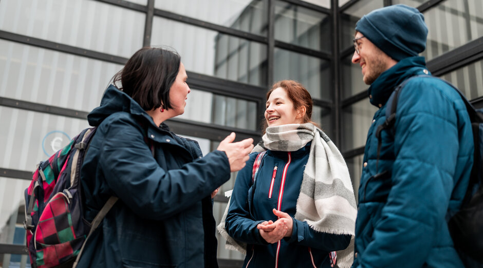 Drei Personen stehen am Campus der Universität Regensburg zusammen und unterhalten sich.