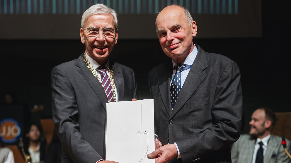 Zwei Herren in dunklen Anzügen lächeln den Betrachter an. Sie halten gemeinsam eine Urkundenmappe hoch.