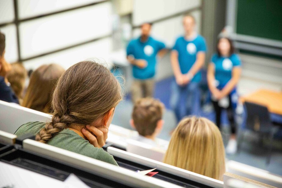 Verschiedene Hinterköpfe in einem Hörsaal sichtbar. Verschwommen im Hintergrund Helferinnen des Schnupperstudiums im blauen T-Shirt erkennbar. (öffnet Vergrößerung des Bildes)