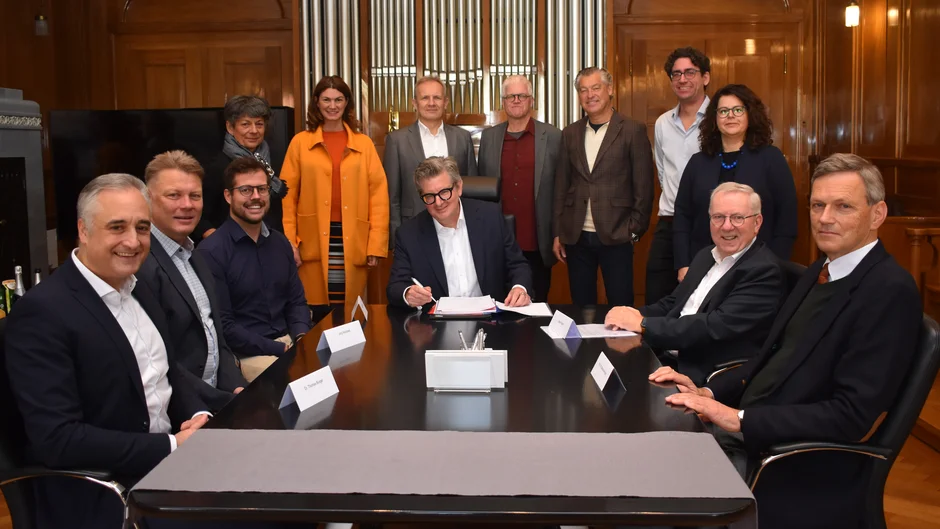 Gruppe von zwölf Personen in einem holzvertäfelten Besprechungsraum. Eine Person sitzt in der Mitte an einem Konferenztisch und unterzeichnet ein Dokument, während die anderen Personen um den Tisch herum sitzen oder stehen. Im Hintergrund sind die Pfeifen einer Orgel zu sehen. (öffnet Vergrößerung des Bildes)