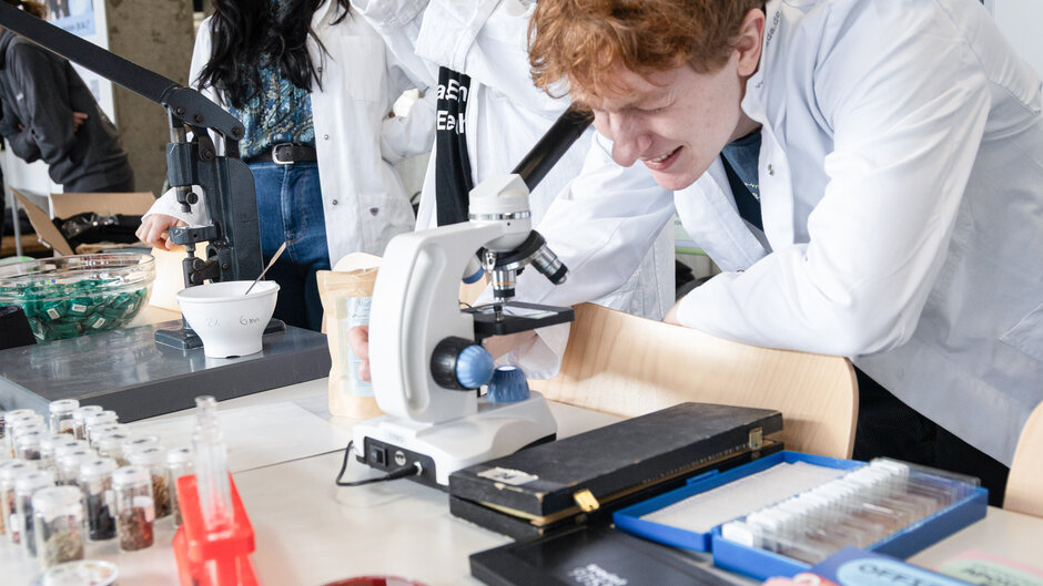 Drei junge Personen in Laborkitteln, eine Frau mit Kopftuch, über Laborgeräte gebeugt. (öffnet Vergrößerung des Bildes)