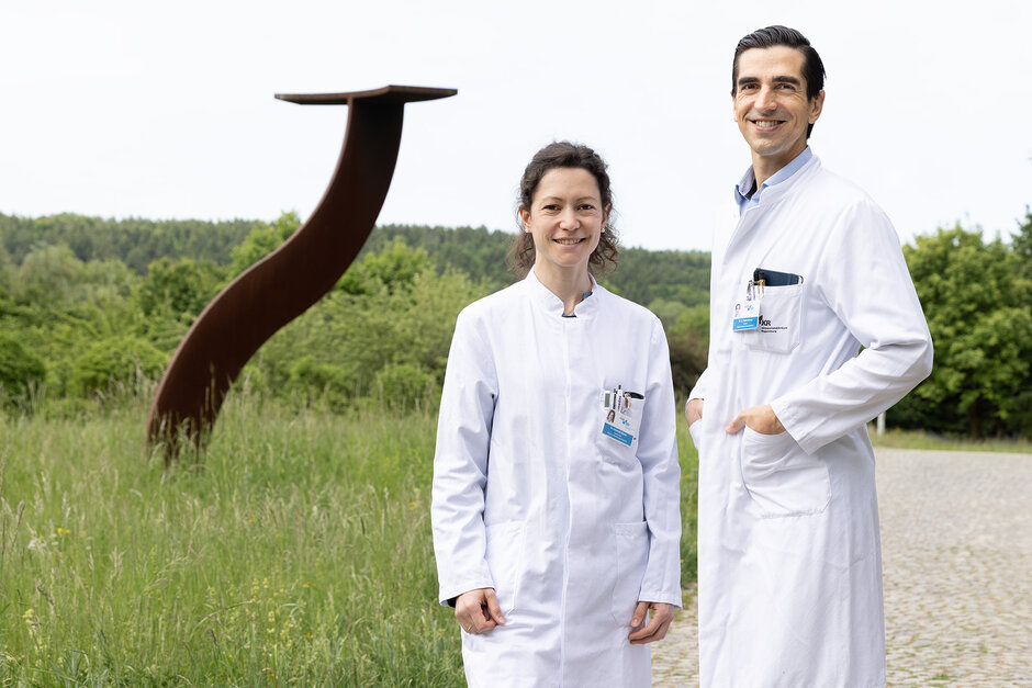 Eine junge Ärztin und ein junger Arzt (beide im weißen Kittel) stehen lächelnd nebeneinander. Das Foto ist im Freien aufgenommen, im Hintergrund sieht man den Teil eines Wegs, ein Stück Wiese und in der Wiese ein Kunstobjekt - eine leicht s-förmig gebogene Säule aus rostig angelaufenem Metall.