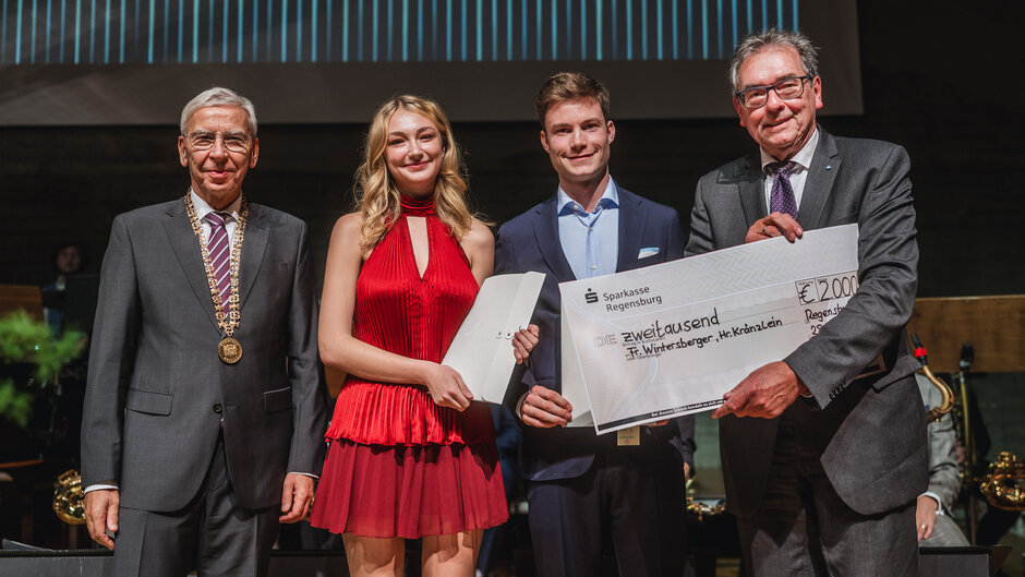 Eine Gruppe aus vier Personen steht nebeneinander auf der Bühne. Von links nach rechts: Ein Herr im dunkelgrauen Anzug mit Krawatte und der Amtskette des Präsidenten der Universität Regensburg um den Hals; eine junge Frau mit langem, blonden Haar und einen roten, kurzen Kleid - sie hält eine Urkundenmappe; ein junger Mann im dunklen Anzug - er hält einen Dekoscheck; ein Herr im dunkelgrauen Anzug mit Brille - er hält den anderen Teil des Dekoschecks.