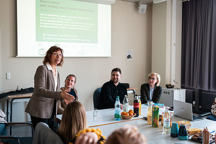 Eine Frau steht in einem Konferenzraum und spricht. Um sie herum sitzen weitere Personen, die ihr aufmerksam zuhören. Im Hintergrund hängt eine Leinwand, auf die eine Präsentation projeziert wird, die Überschrift lautet "Meet & Greet". (öffnet Vergrößerung des Bildes)