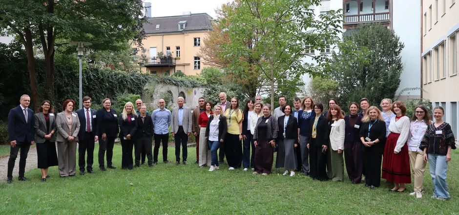 Große Gruppe von Teilnehmenden steht für ein Gruppenfoto auf einer Rasenfläche in einem begrünten Innenhof. Im Hintergrund sind Bäume, Sträucher und mehrstöckige Wohngebäude zu sehen. Einige Personen tragen Namensschilder, und die Gruppe ist in einer Reihe für die Aufnahme aufgestellt. (öffnet Vergrößerung des Bildes)