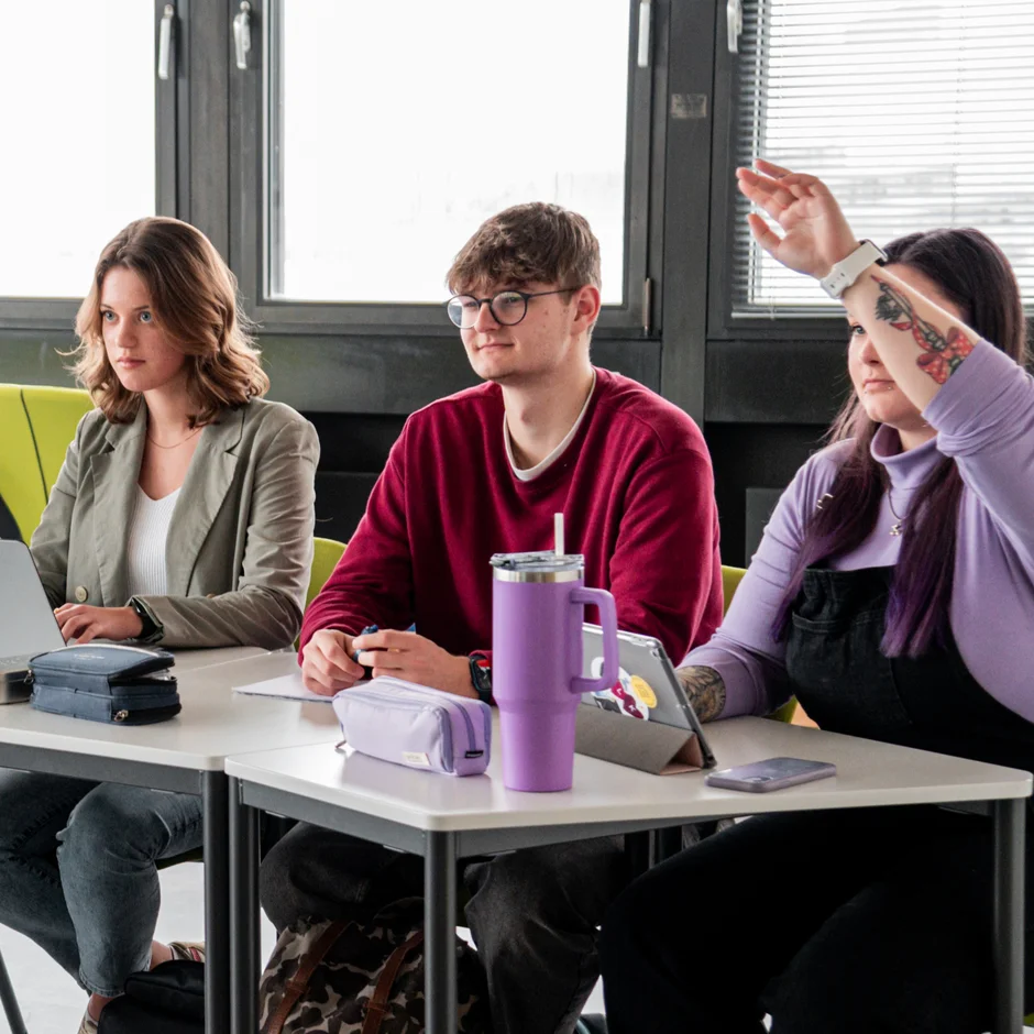Studierende an einem Tisch in einem Seminarraum. Zwei Studentinnen melden sich.
