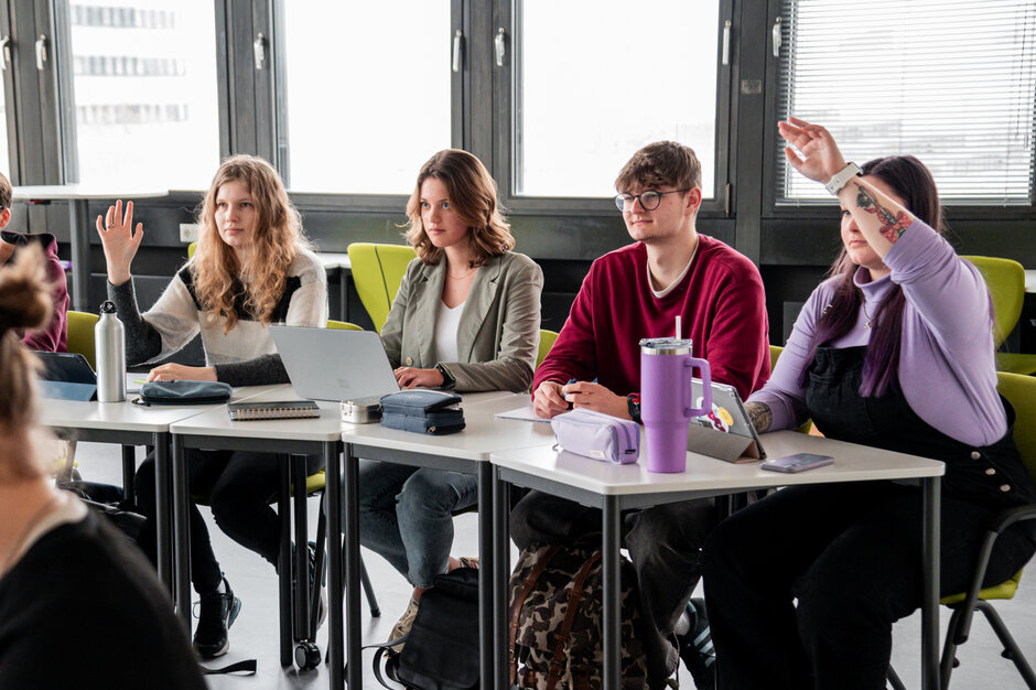 Studierende an einem Tisch in einem Seminarraum. Zwei Studentinnen melden sich.
