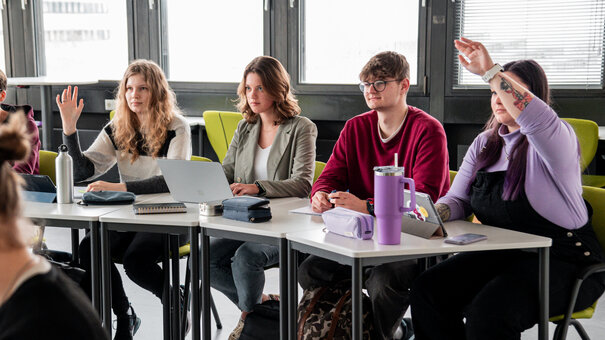 Studierende an einem Tisch in einem Seminarraum. Zwei Studentinnen melden sich.