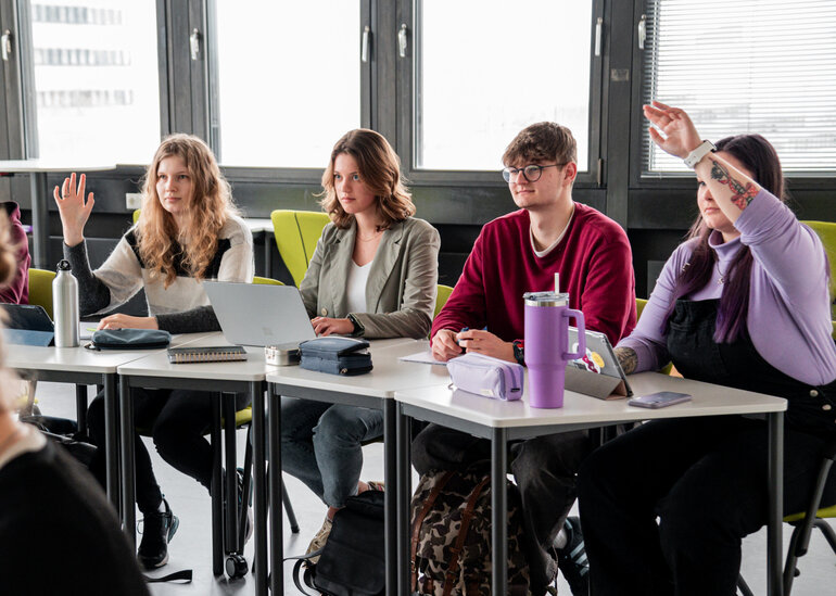 Studierende an einem Tisch in einem Seminarraum. Zwei Studentinnen melden sich.