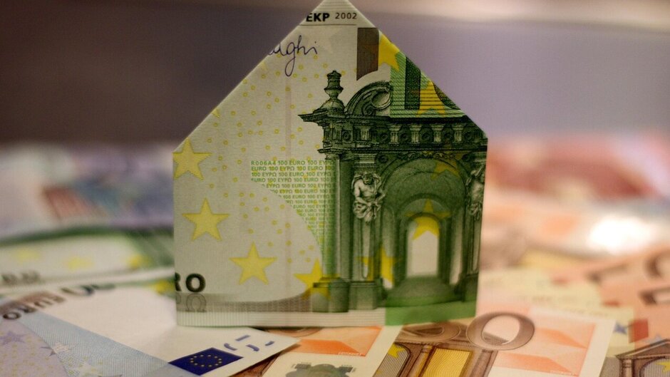 Ein Geldschein in Hausform gefaltet steht auf anderen Geldscheinen