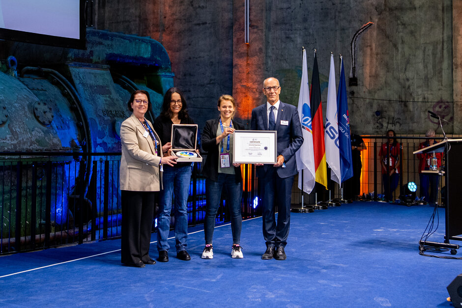 Vier Personen, drei Frauen und ein Mann, bei der Übergabe von zwei Urkunden. Sie lächeln alle in die Kamera. (öffnet Vergrößerung des Bildes)
