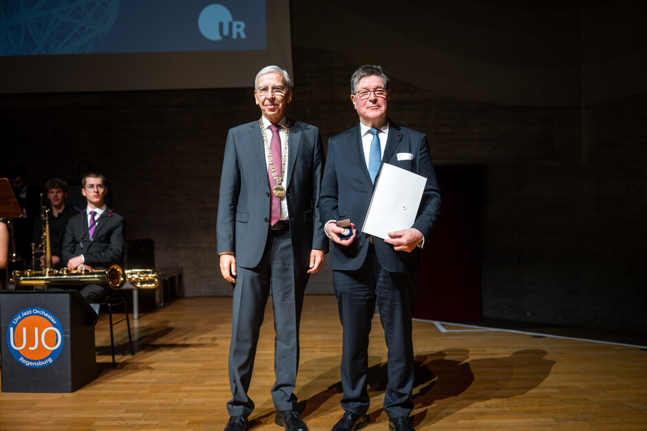 Zwei Personen stehen auf einer Bühne und posieren für ein Foto. Die Person rechts hält eine Urkundenmappe und ein kleines Etui in den Händen. Die Person links trägt einen Anzug mit einer Amtskette. Im Hintergrund sitzen Mitglieder eines Blasensembles.