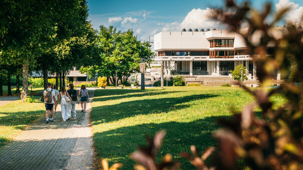 Eine Gruppe Studierender geht über den Campus der Universität Regensburg. Im Hintergrund ist die Zentralbibliothek zu sehen.