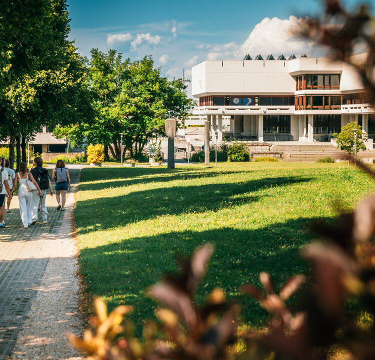 Eine Gruppe Studierender geht über den Campus der Universität Regensburg. Im Hintergrund ist die Zentralbibliothek zu sehen.