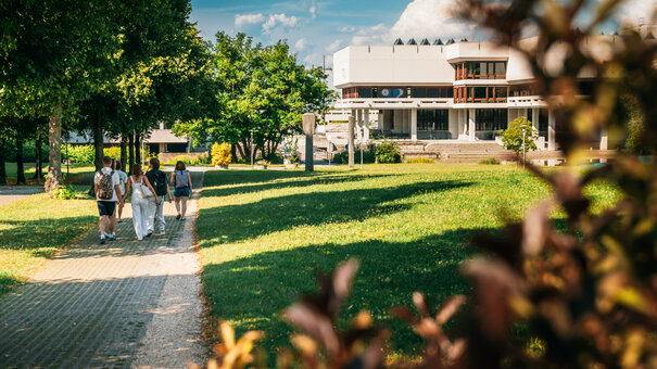 Eine Gruppe Studierender geht über den Campus der Universität Regensburg. Im Hintergrund ist die Zentralbibliothek zu sehen.