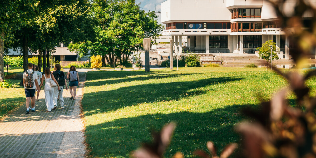 Eine Gruppe Studierender geht über den Campus der Universität Regensburg. Im Hintergrund ist die Zentralbibliothek zu sehen.