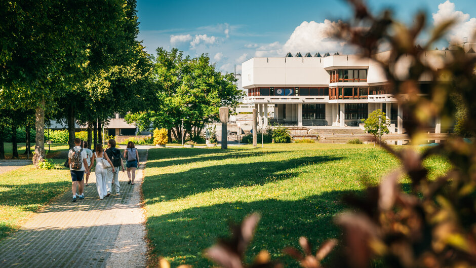 Eine Gruppe Studierender geht über den Campus der Universität Regensburg. Im Hintergrund ist die Zentralbibliothek zu sehen.