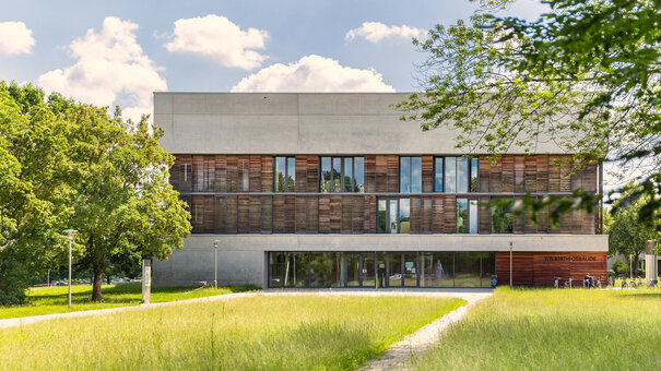 Ein Hochschulgebäude mit Beton- und Holzfassade steht in einer parkähnlichen Umgebung umgeben von grüner Wiese, grünen Bäumen. Die Sonne scheint, der Himmel ist blau mit vereinzelten weißen Wolken.