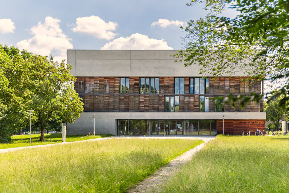 Ein Hochschulgebäude mit Beton- und Holzfassade steht in einer parkähnlichen Umgebung umgeben von grüner Wiese, grünen Bäumen. Die Sonne scheint, der Himmel ist blau mit vereinzelten weißen Wolken.