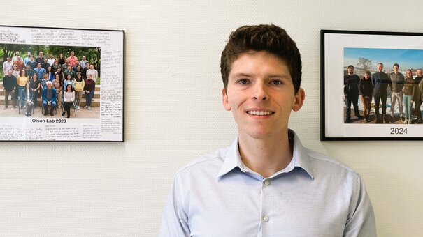 Junger Mann lächelnd, im hellen Hemd, stehend vor Wand mit gerahmten Gruppenfotos hinter sich.