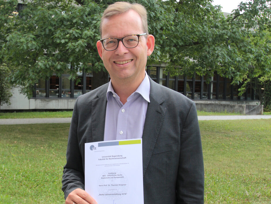 Prof. Kingreen steht lächelnd auf der Freifläche vor dem Vielberthgebäude und hält die Urkunde zum Lehrpreis in den Händen. (öffnet Vergrößerung des Bildes)