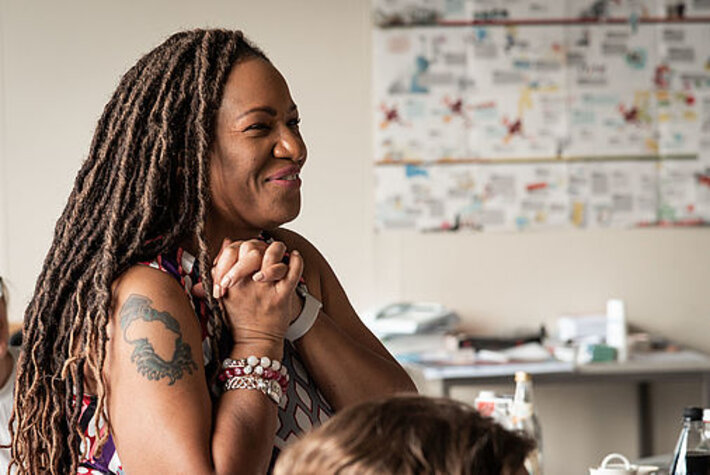 Eine Frau im Halbprofil mit einander verschränkten Händen, die sie an ihren Oberkörper hält. Sie lächelt. Auf ihrem Oberarm ist ein Tatoo mit dem Umriss des afrikanischen Kontinents zu erkennen. (öffnet Vergrößerung des Bildes)