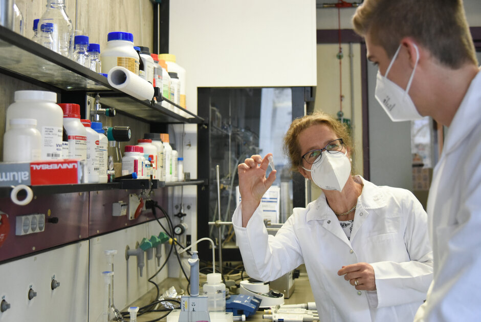 Zwei Personen mit FFP2-Masken und Laborkitteln stehen in einem Labor, umgeben von Regalen, auf denen Chemikalien aufgereiht sind. (öffnet Vergrößerung des Bildes)