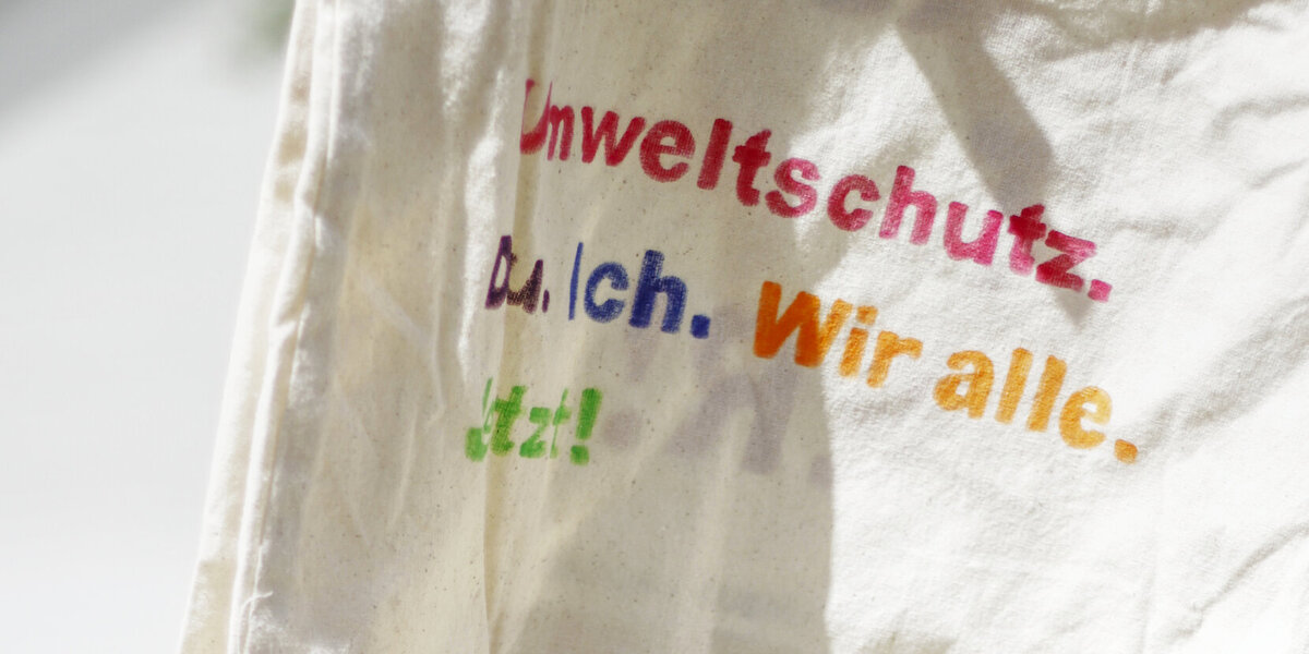Auf einer natur-weißen Stofftasche steht in bunten Farben: "Umweltschutz. Du. Ich. Wir alle. Jetzt!" Im Hintergrund ist eine Gruppe junger Menschen. 