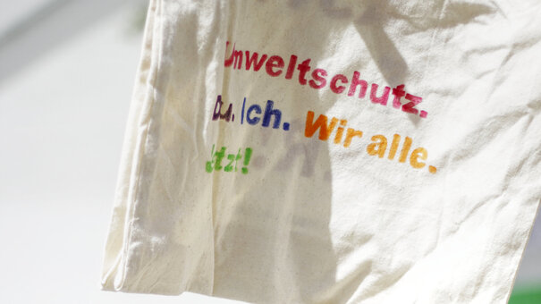 Auf einer natur-weißen Stofftasche steht in bunten Farben: "Umweltschutz. Du. Ich. Wir alle. Jetzt!" Im Hintergrund ist eine Gruppe junger Menschen. 