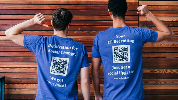 Zwei Personen stehen mit dem Rücken zum Betrachter vor einer holzvertäfelten Wand. Den jeweils äußeren Arm haben sie erhoben, der Ellbogen ist jeweils abgewinkelt und mit dem jeweiligen Daumen zeigen sie auf ihre T-Shirts. Dort sind Hinweise auf "VoluLink" und QR-Codes aufgedruckt.
