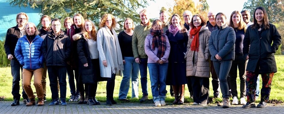 Etwa 20 Personen stehen für ein Gruppenfoto im Freien auf einem Weg vor einer Wiese und Bäumen, gekleidet in winterliche Jacken und Mäntel bei sonnigem Wetter. (öffnet Vergrößerung des Bildes)