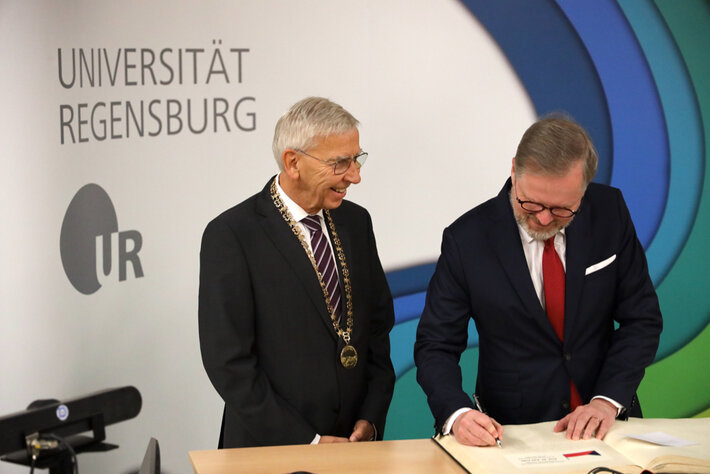 Zwei Männer in formeller Kleidung. Die links stehende Person trägt eine Amtskette. Die rechts stehende Person schreibt in ein großformatiges Buch; auf der Seite ist die tschechische Flagge zu erkennen. Im Hintergund ist ein Aufsteller mit der Aufschrift "Universität Regensburg" und dem Logo der Universität zu sehen. (öffnet Vergrößerung des Bildes)