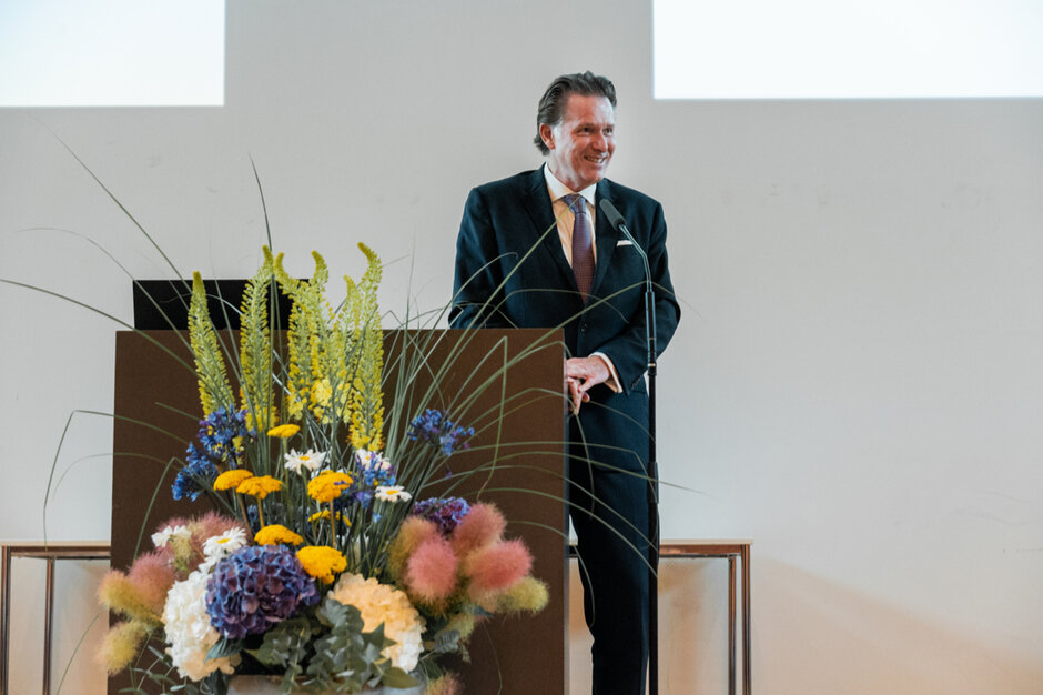 Ein Mann steht seitlich an einem Rednerpult und spricht lächelnd. Vor dem Rednerpult steht ein üppiger Blumenschmuck.