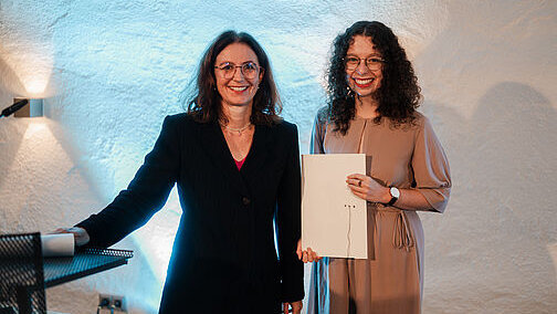 Zwei dunkelhaarige Frauen mit Brille - die eine schwarz, die andere beige gekleidet - lächeln in die Kamera. Die rechts stehende junge Frau in beigem Kleid hält eine Urkundenmappe in der Hand.