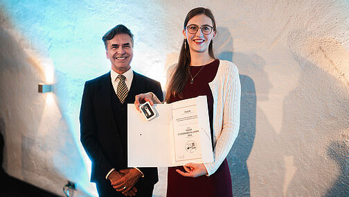 Ein Herr im dunklen Dreiteiler steht neben einer jungen Frau mit Brille und langen, zusammengebundenen Haaren, die eine aufgeschlagene Urkundenmappe mit dem Inhalt zum Betrachter hält. 