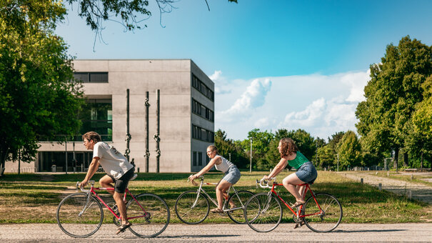 Drei Studierende fahren mit ihren Fahrrädern über den sommerlichen Campus der Universität Regensburg.