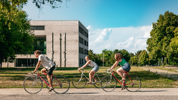 Drei Studierende fahren mit ihren Fahrrädern über den sommerlichen Campus der Universität Regensburg.