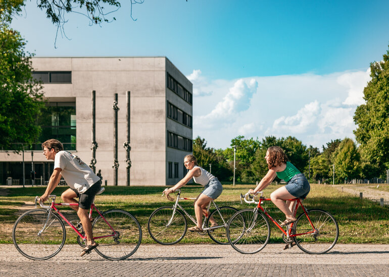 Drei Studierende fahren mit ihren Fahrrädern über den sommerlichen Campus der Universität Regensburg.