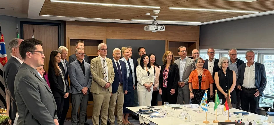 Gruppenfoto einer internationalen Hochschuldelegation in einem modern ausgestatteten Besprechungsraum. Die Teilnehmenden stehen hinter einem Konferenztisch mit Unterlagen, Kaffeetassen und Tischflaggen. Das Bild entstand im Rahmen eines gemeinsamen Austauschs beziehungsweise einer Kooperation zwischen Hochschulen. (öffnet Vergrößerung des Bildes)