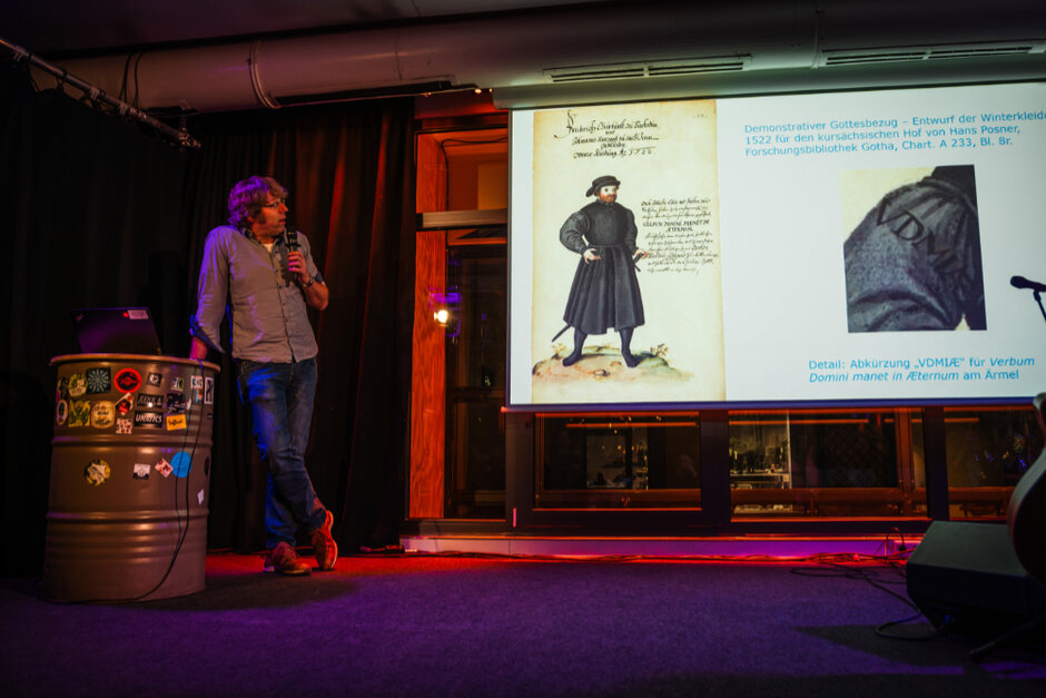 Ein Mann steht auf einer Bühne, hält ein Mikrophon in der Hand und blickt zu einer Projektion auf einer Leinwand.