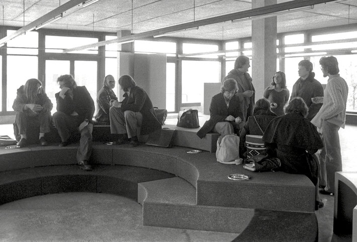 Das schwarz-weiß Foto zeigt junge Männer und Frauen, die sich um eine Sitzlandschaft herum gruppieren. Frisuren und Kleidung entsprechen der Mode der 1970er Jahre. (opens enlarged image)
