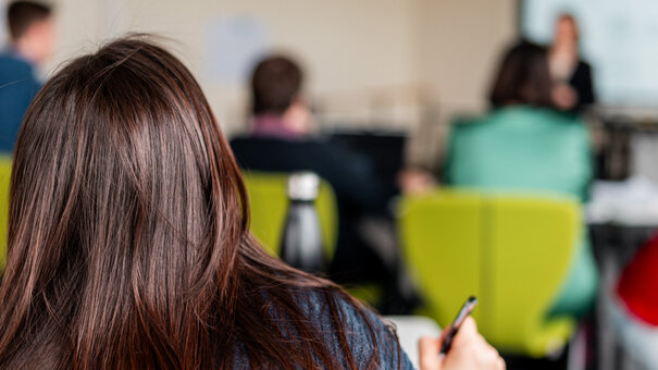 Eine Studentin arbeitet an einem Tisch sitzend. Im Hintergrund sind weitere Studierende und eine Dozentin zu sehen.