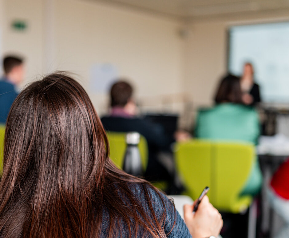Eine Studentin arbeitet an einem Tisch sitzend. Im Hintergrund sind weitere Studierende und eine Dozentin zu sehen.