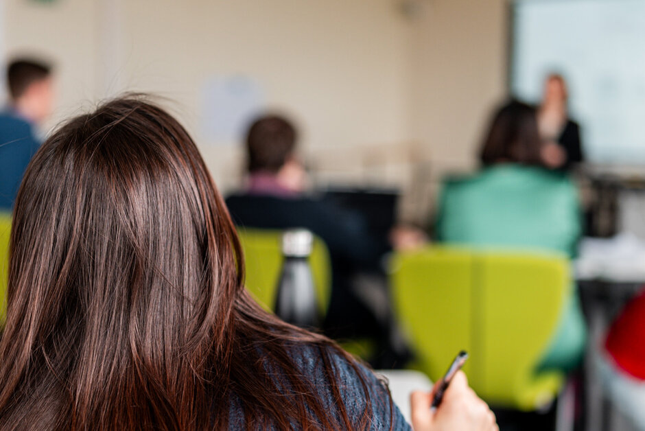 Eine Studentin arbeitet an einem Tisch sitzend. Im Hintergrund sind weitere Studierende und eine Dozentin zu sehen.