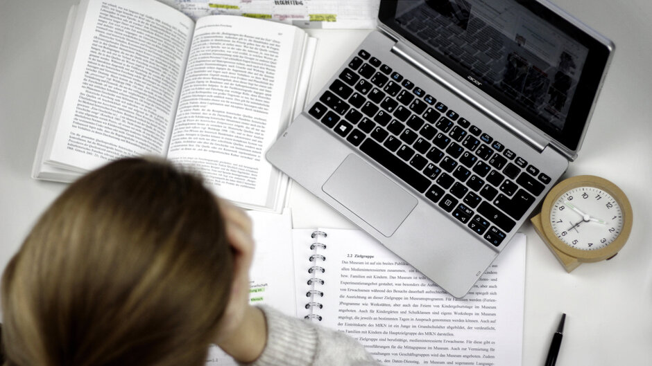 Eine Studentin sitzt an einem Schreibtisch, auf dem ein Laptop, ein Wecker, auf aufgeschlagenes Buch und ein Vorlesungsskript liegen.