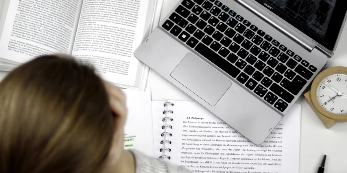Eine Studentin sitzt an einem Schreibtisch, auf dem ein Laptop, ein Wecker, auf aufgeschlagenes Buch und ein Vorlesungsskript liegen.