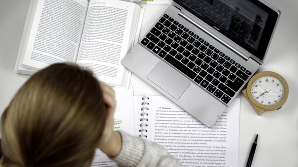 Eine Studentin sitzt an einem Schreibtisch, auf dem ein Laptop, ein Wecker, auf aufgeschlagenes Buch und ein Vorlesungsskript liegen.