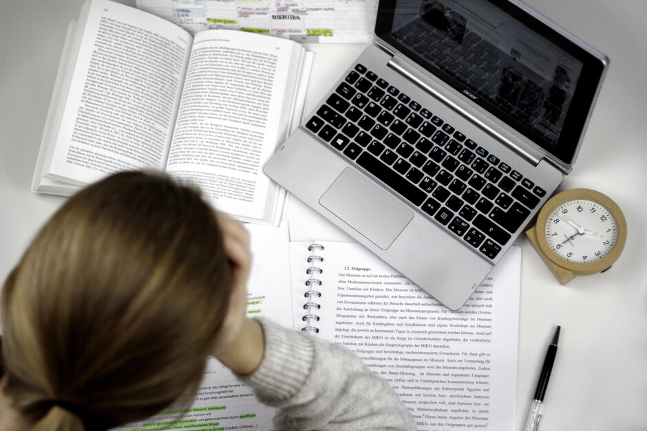 Eine Studentin sitzt an einem Schreibtisch, auf dem ein Laptop, ein Wecker, auf aufgeschlagenes Buch und ein Vorlesungsskript liegen.