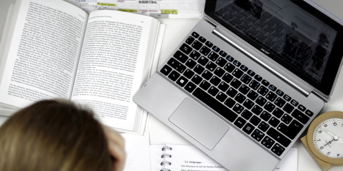 Eine Studentin sitzt an einem Schreibtisch, auf dem ein Laptop, ein Wecker, auf aufgeschlagenes Buch und ein Vorlesungsskript liegen.