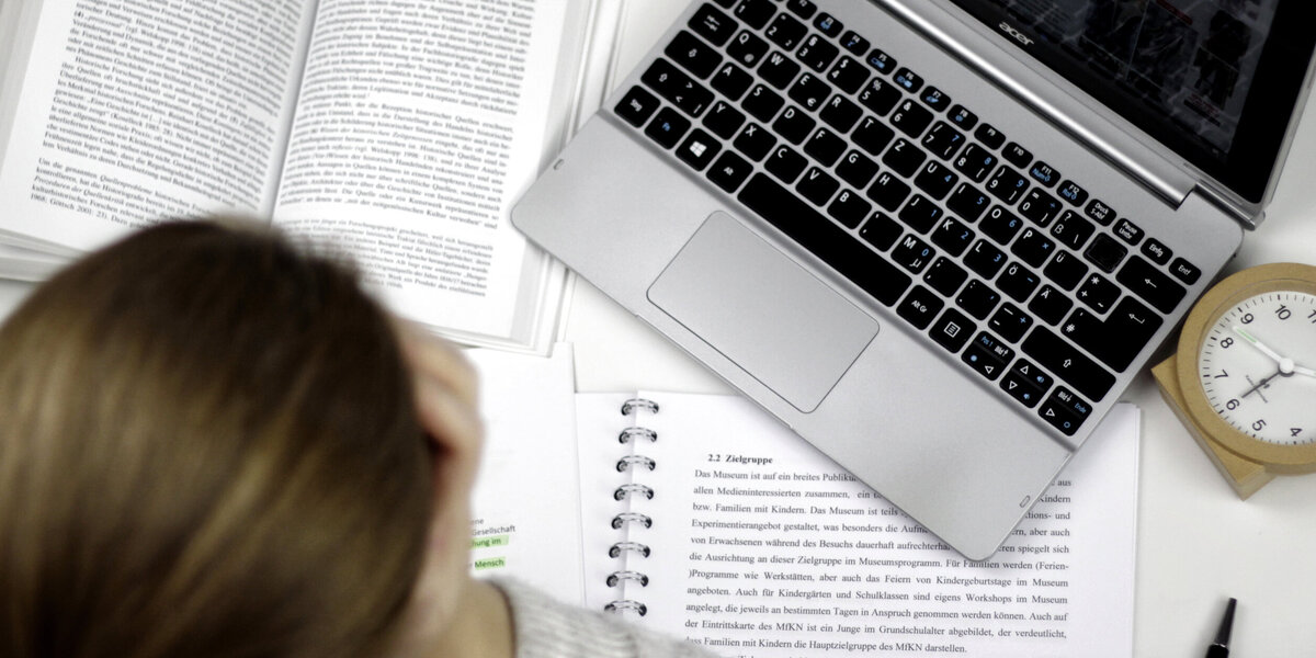 Eine Studentin sitzt an einem Schreibtisch, auf dem ein Laptop, ein Wecker, auf aufgeschlagenes Buch und ein Vorlesungsskript liegen.